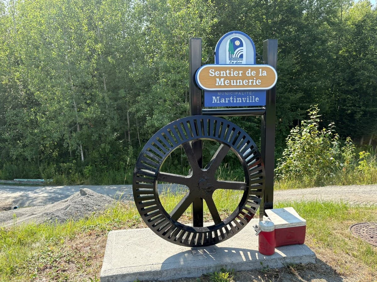 Panneau du Sentier de la Meunerie et grande roue dentée de moulin posée sur une dalle de béton