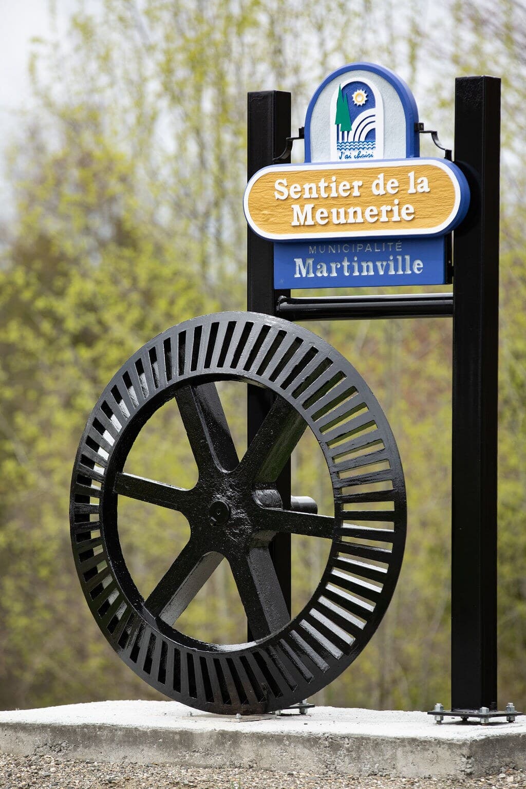 Le panneau du Sentier de la Meunerie et sa roue de moulin en fonte, sur son socle